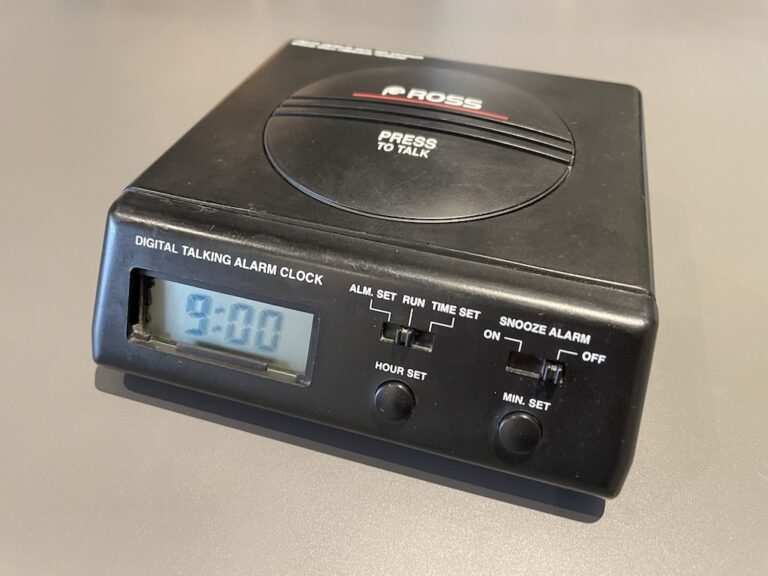 A black alarm clock made by Ross with text on the front that says 'digital talking alarm clock', a large circular button on top labelled 'Press to talk', and the time displayed on a digital display on the front alongside buttons for setting the time and alarm